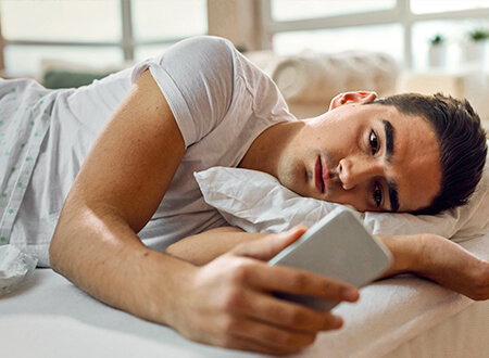 Mann liegt im Bett und schaut traurig auf sein Smartphone