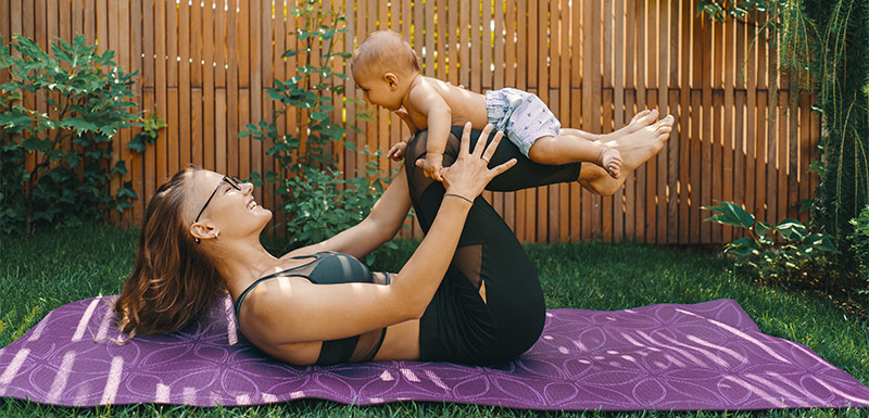 Junge Mutter praktiziert Yoga in Rückenlage mit Ihrem Säugling auf den Schienbeinen liegend.